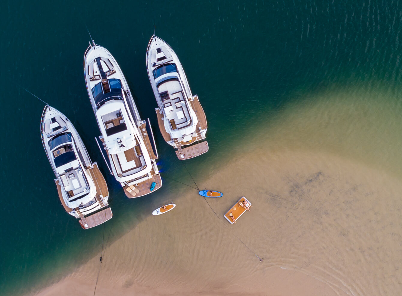 Three Galeon yachts anchored in the water with paddleboards behind them near the sand