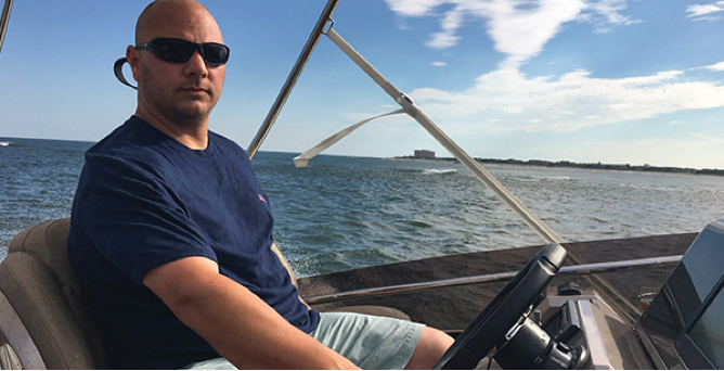Man with dark sunglasses and a navy t-shirt at the driver's seat of a boat