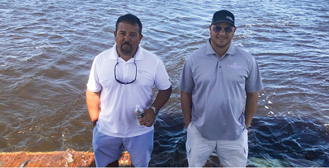 Two men standing by the water with their hands in their pockets, one with a hat on