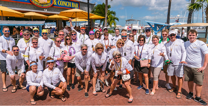 A large group of men and women smiling in rows in front of a building with yellow umbrellas outside