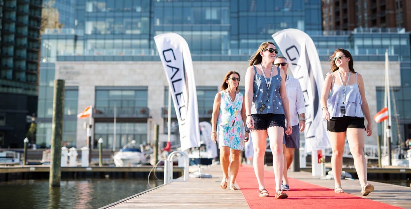 Four people walking down a walkway with a red carpet and Galeon flags in front of the building behind them