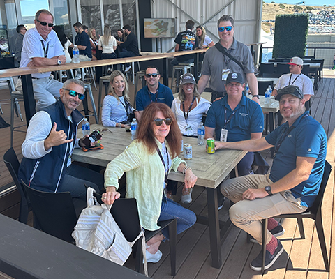 Group of nine people smiling and sitting at an outdoor wooden deck during a sunny day.