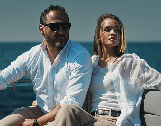 A man in sunglasses and a woman with flowing hair sit in front of a calm sea.